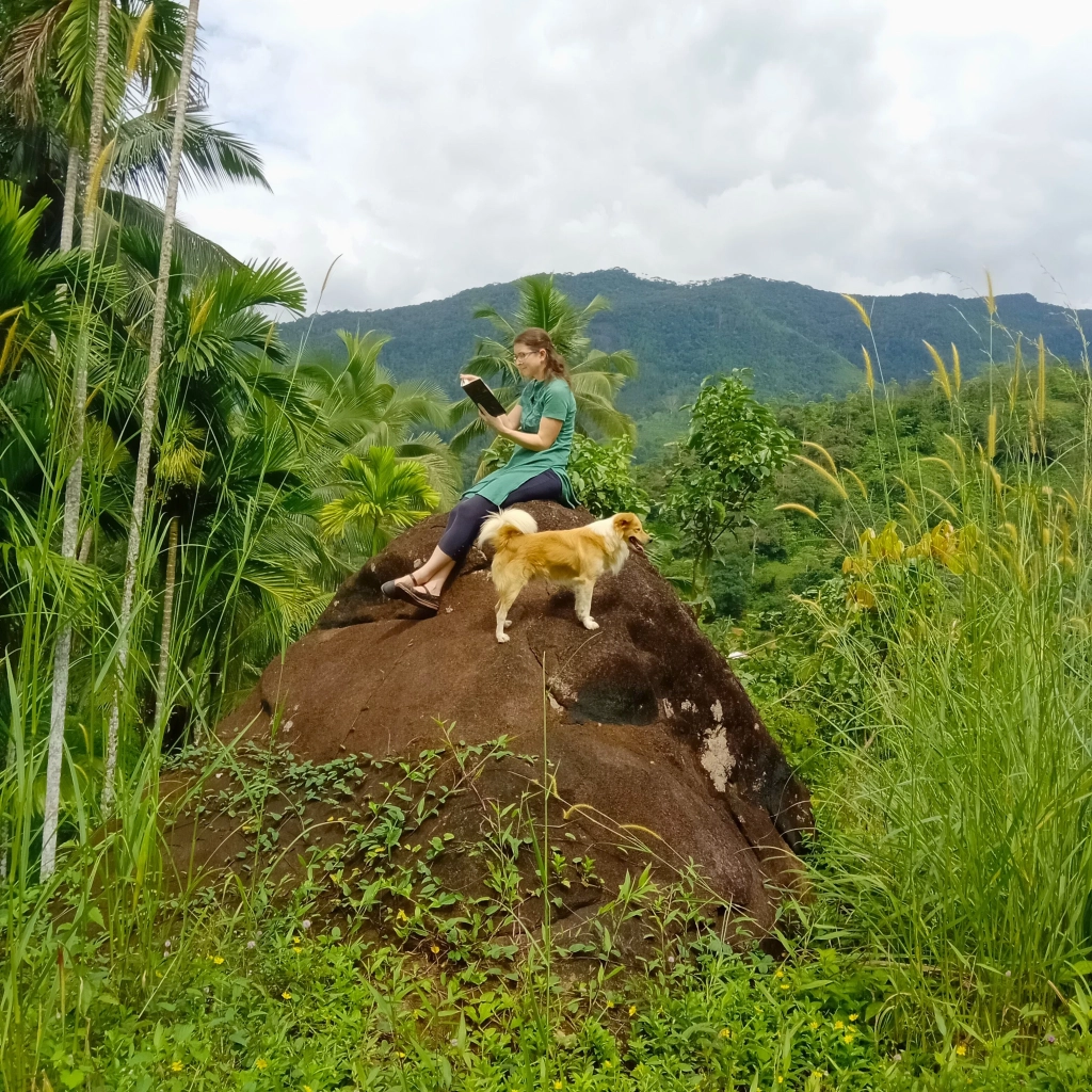 Joanne reading a book on a big rock surrounded by tropical greenery, with a dog.